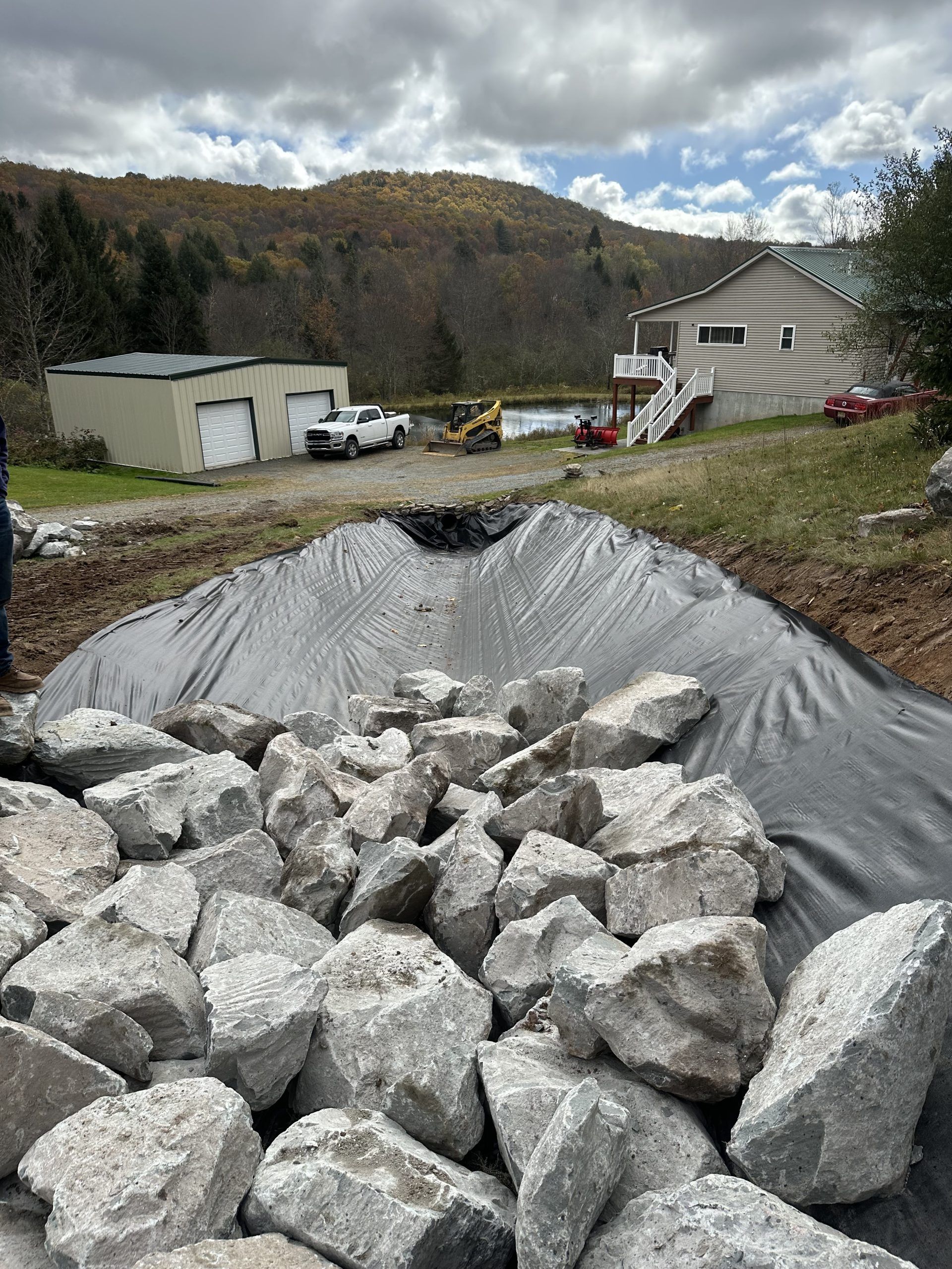 Rocks piled in front of a black liner, with a house, vehicles, and fall foliage visible in the background.