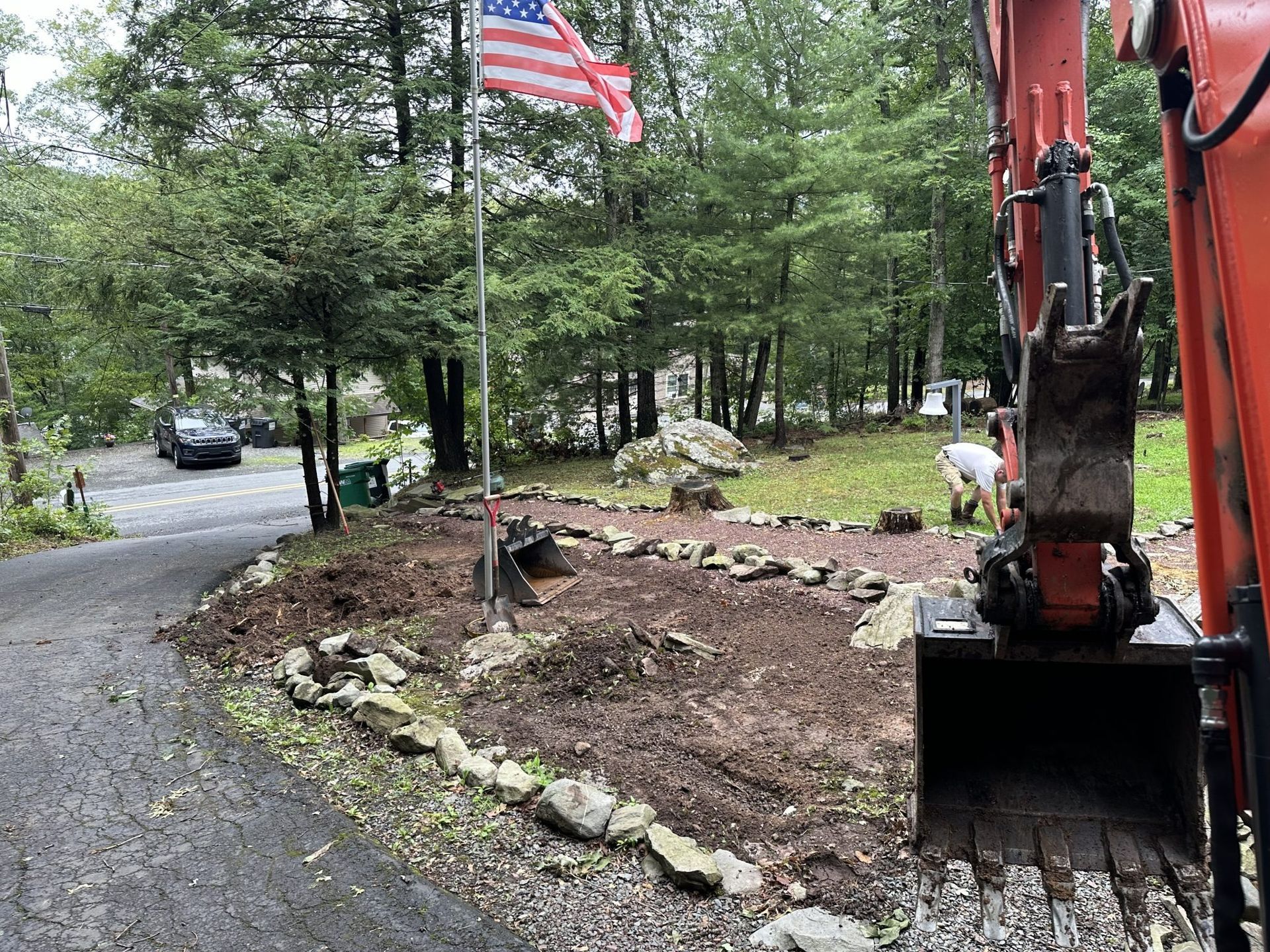 An excavator and person working on a landscaped area with rocks, dirt, and an American flag.