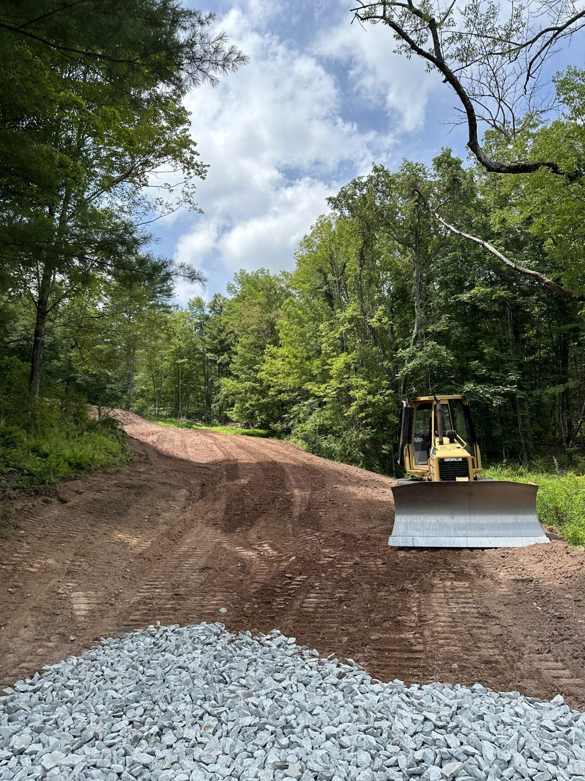 Bulldozer on a dirt road being constructed through a wooded area. The sky is partly cloudy.