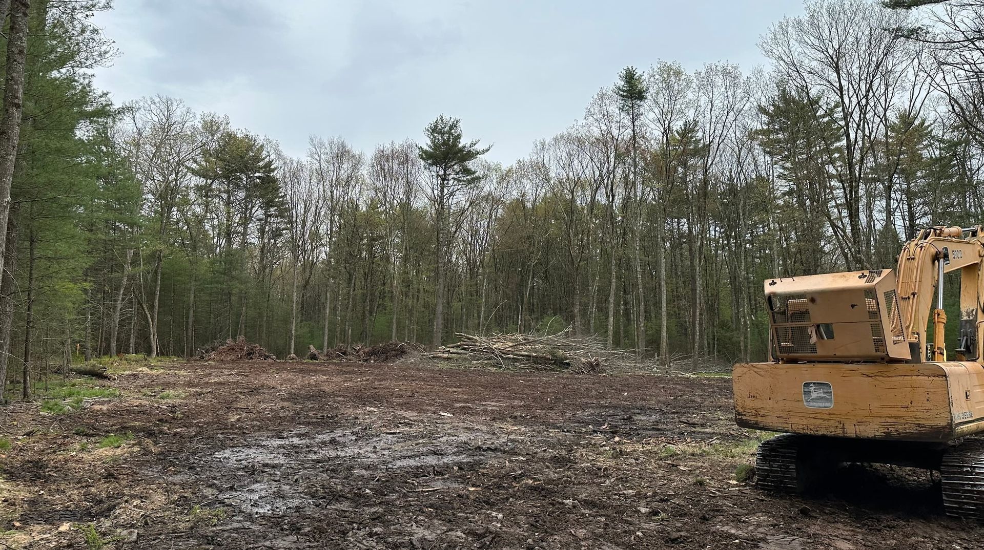 Cleared land with a yellow excavator, trees in the background, overcast sky.