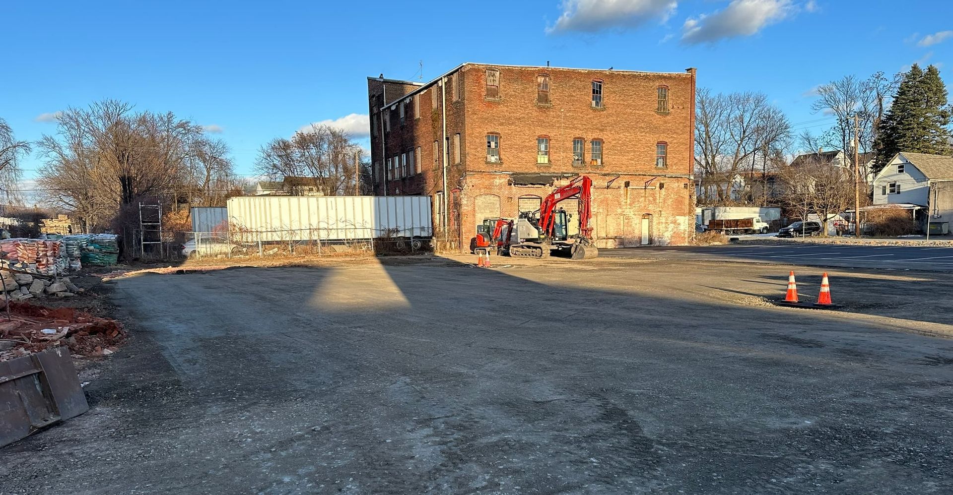 Remains of brick building, construction site, excavator. Open lot, clear sky, two orange traffic cones.