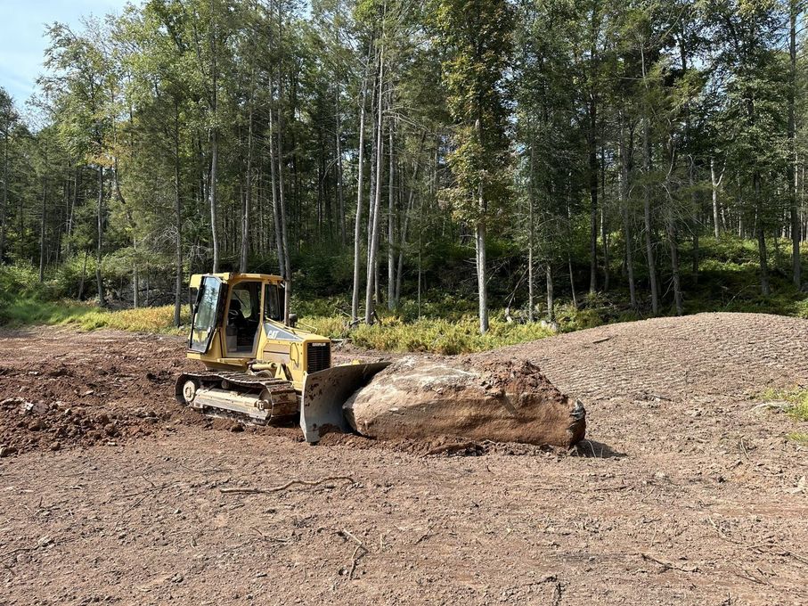 Yellow bulldozer pushing a large mound of dirt in a clearing; trees in background.