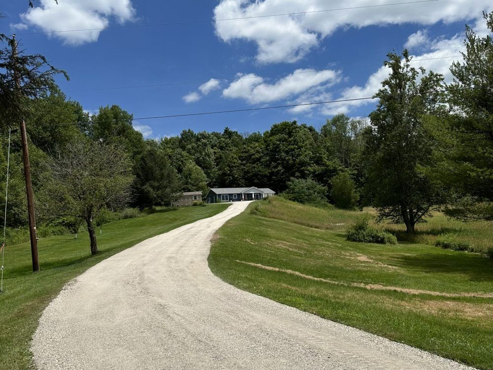 Gravel driveway curves uphill to a white ranch house surrounded by trees under a blue sky with clouds.