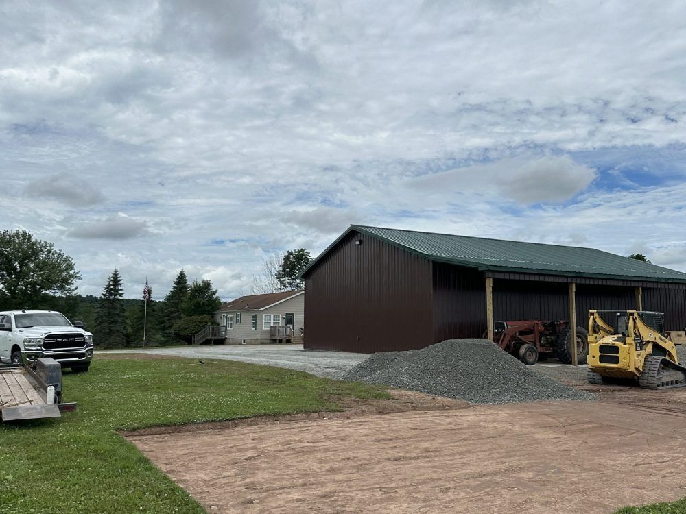 Construction site with brown building, gravel pile, and yellow loader. White truck with trailer on grass.