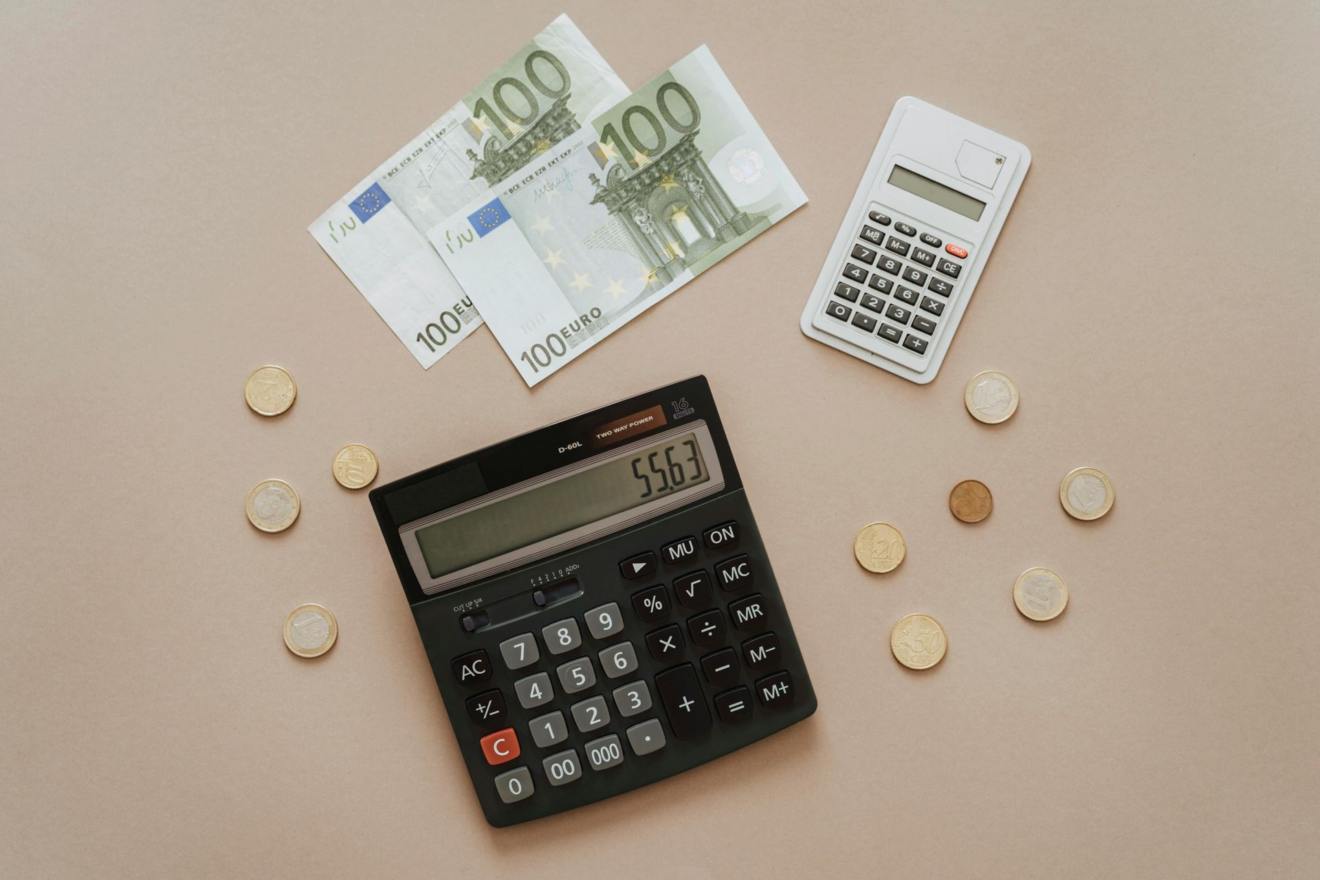 Two calculators, two 100 Euro bills, and coins on a neutral surface, implying financial calculations.