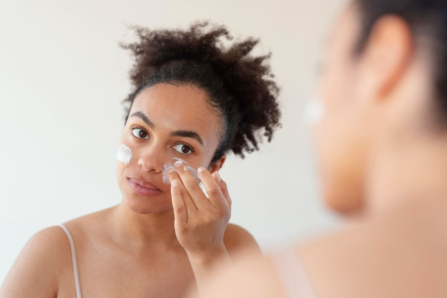 Woman with curly hair applying cream to her face in front of a mirror, indoors.