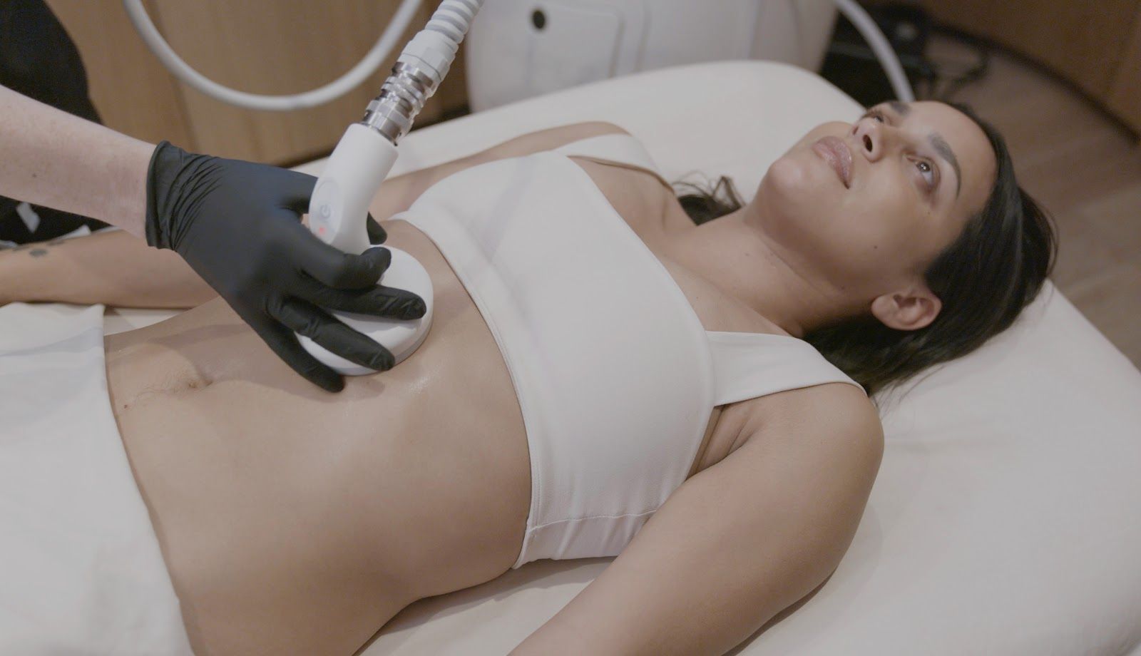 A practitioner in black gloves uses a cosmetic device on a person's abdomen during a body contouring procedure.