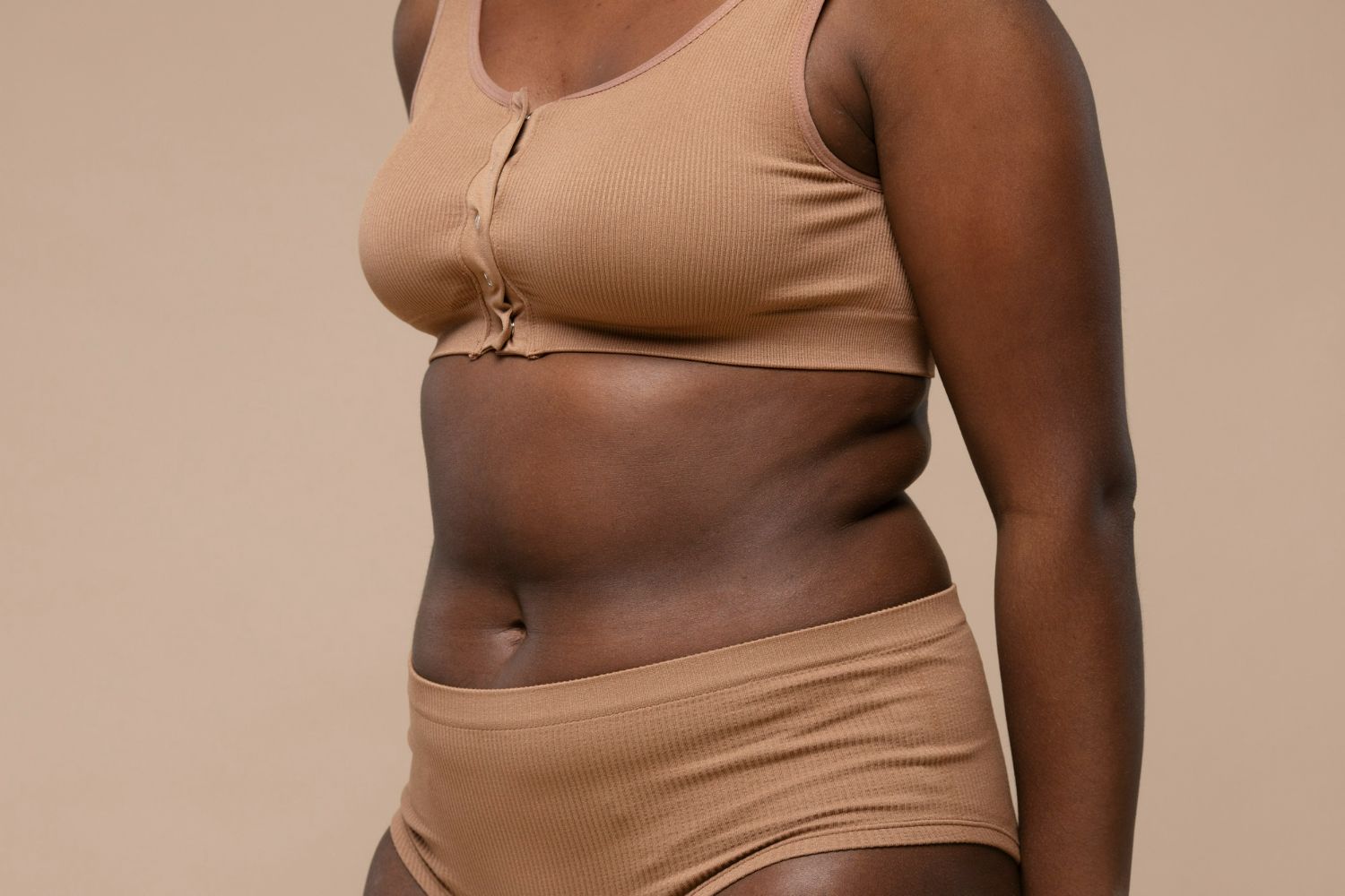 Woman with dark skin wearing a tan ribbed bra and shorts, against a tan backdrop.