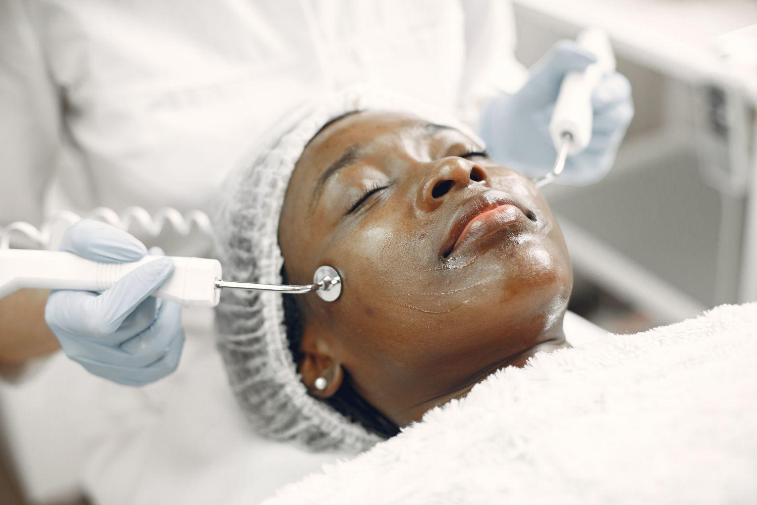Black woman receiving facial treatment with instruments, in a white medical setting.