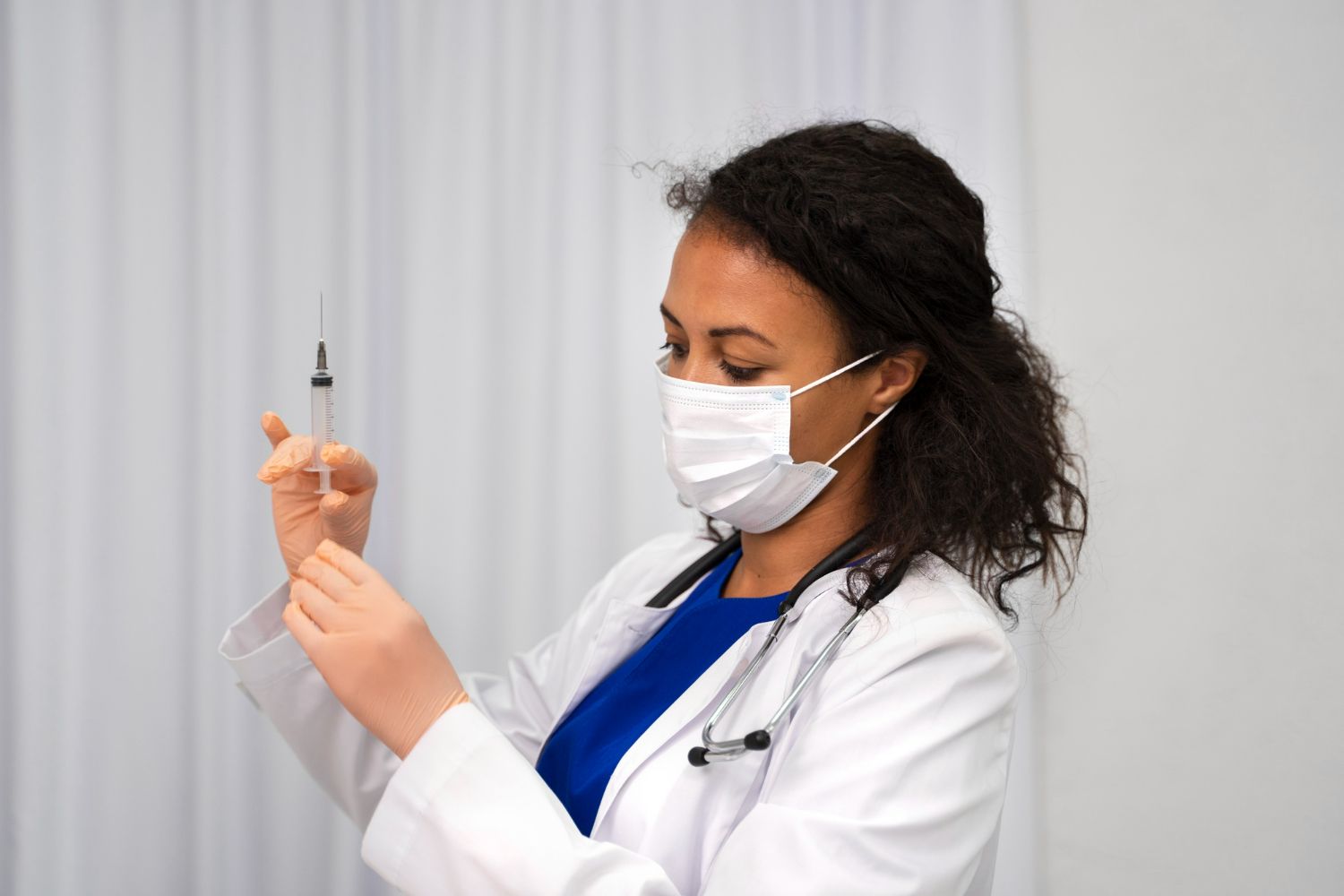Female doctor in mask preparing syringe. White coat, stethoscope, white background.