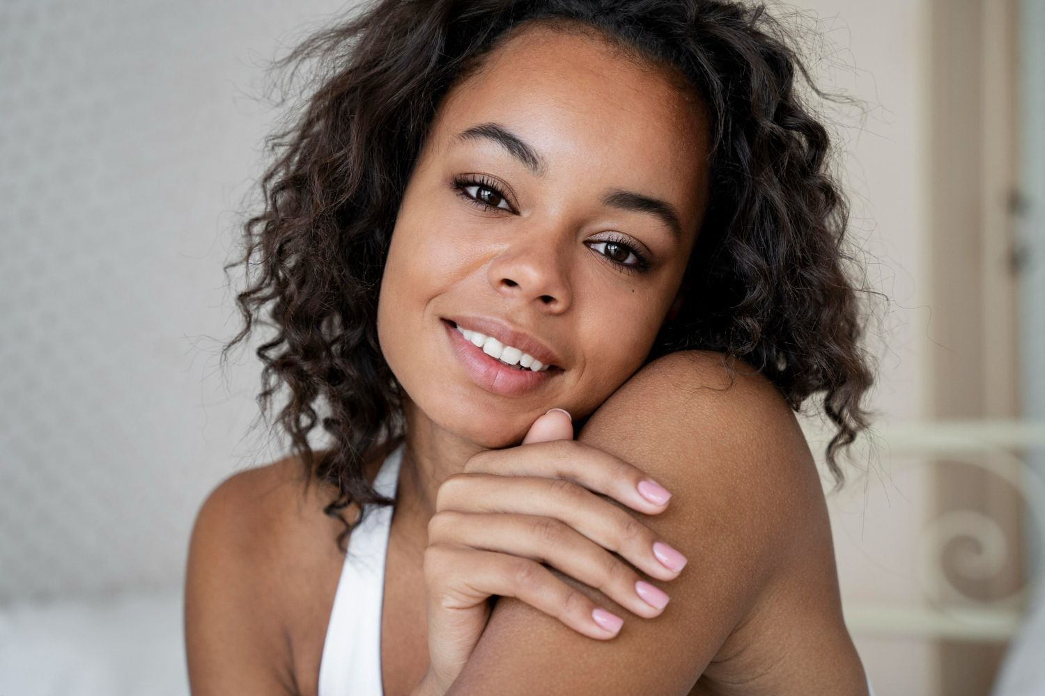 Smiling Black woman with curly hair, in white top, resting chin on shoulder.