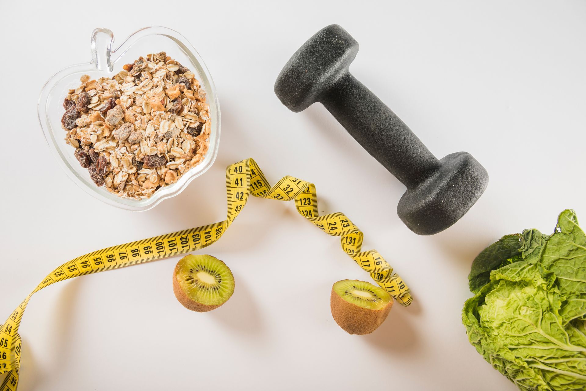 A bowl of granola , a dumbbell , kiwi , and a measuring tape on a table.