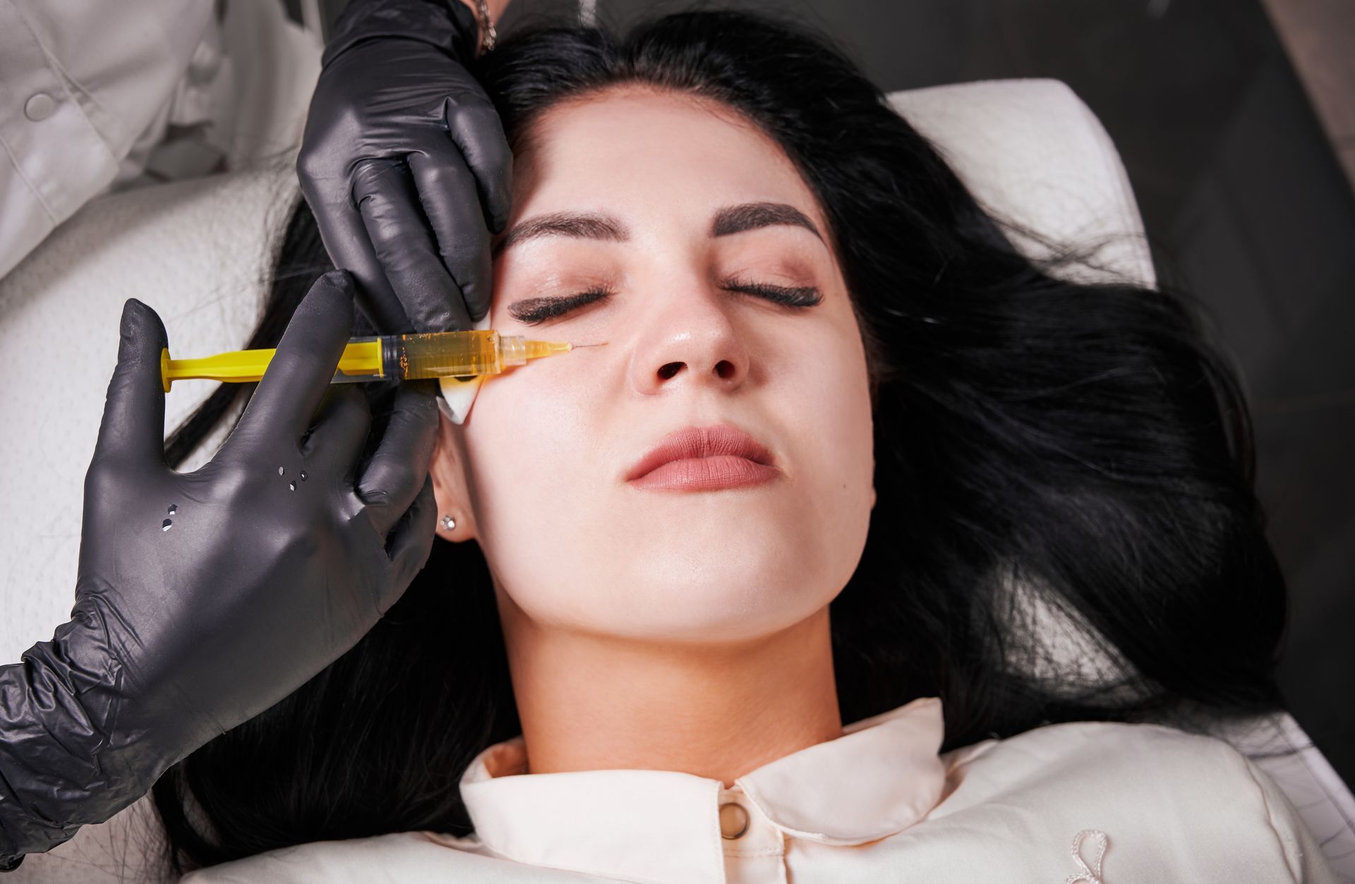 Woman receiving a facial injection near the eye, hands in black gloves hold a syringe.