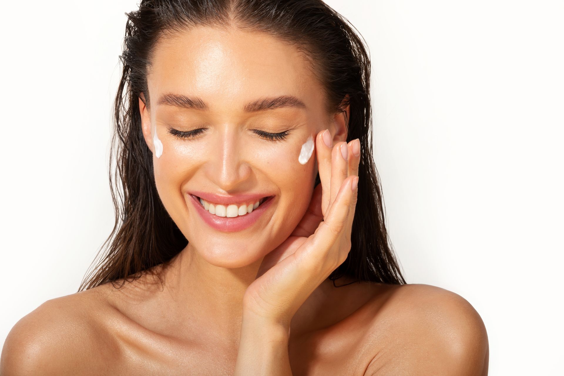 A woman is smiling while applying cream to her face.