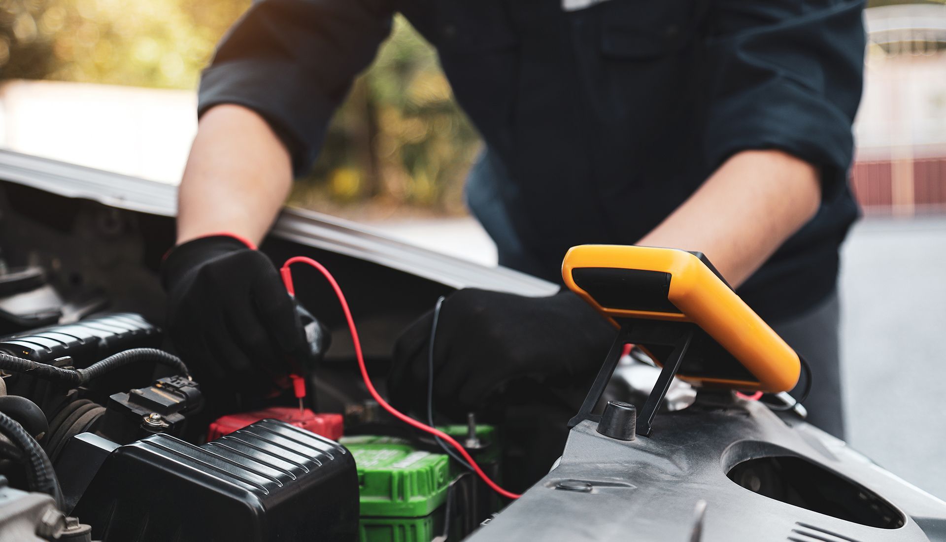 Mechanic tests a car battery with a multimeter under the hood. Black gloves and red test leads.