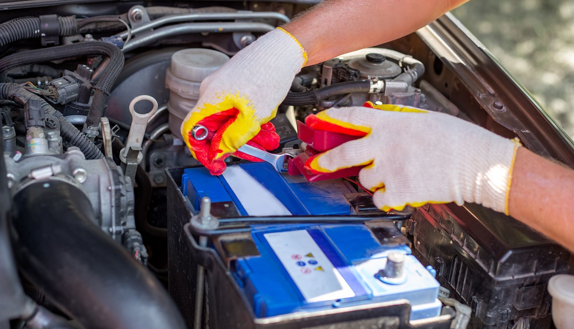 Hands wearing gloves working on a car battery in an engine bay.