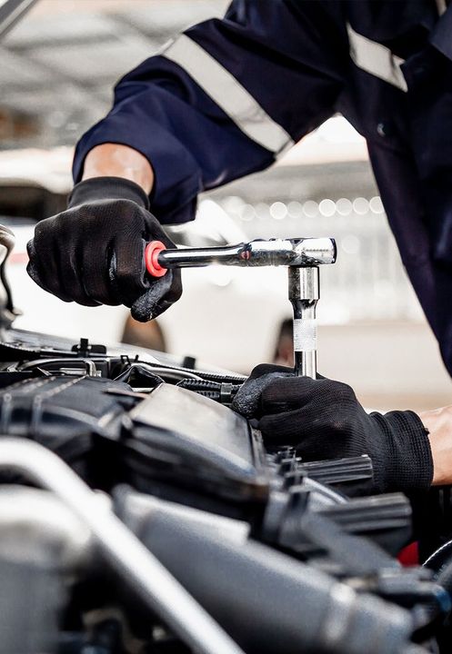Mechanic in blue jumpsuit using a wrench to work on a car engine, wearing black gloves.