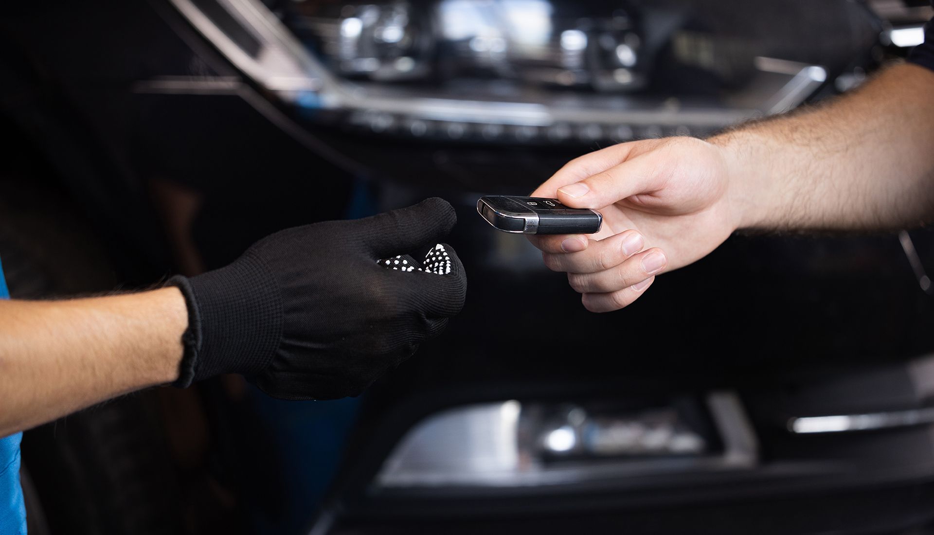Hands exchanging car keys in front of a black car. One hand wears a black glove.