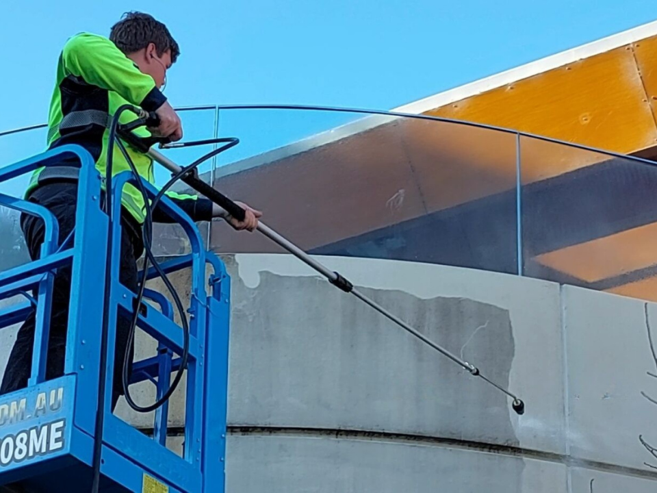 A worker pressure washing a concrete wall from a blue lift; shows
