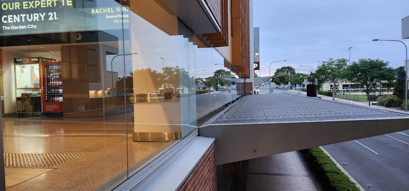 View of a building's glass entrance with a metal awning extending over a sidewalk and road at dusk.