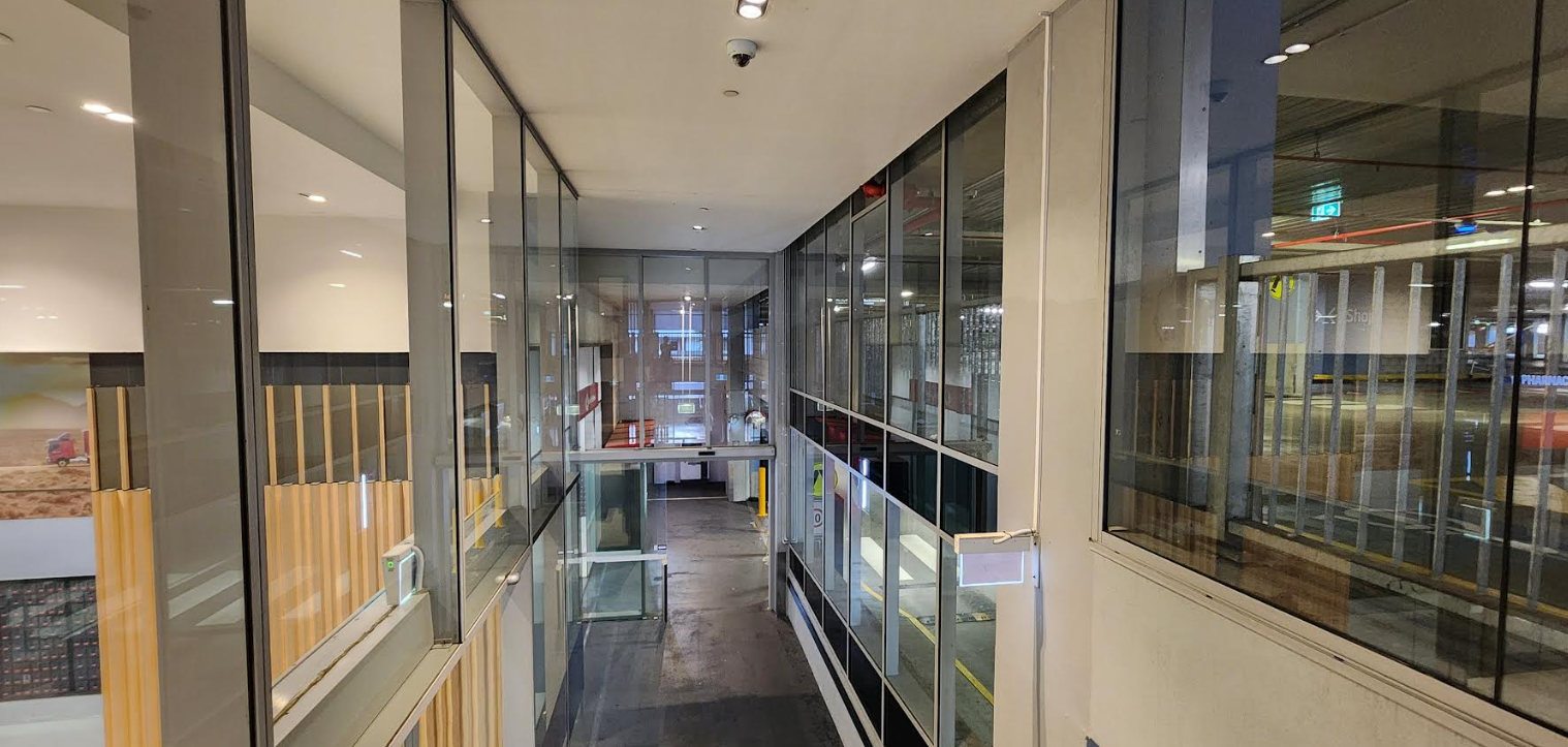 A narrow corridor between glass walls, with wooden slatted accents on the left and a view into a lit room at the end.