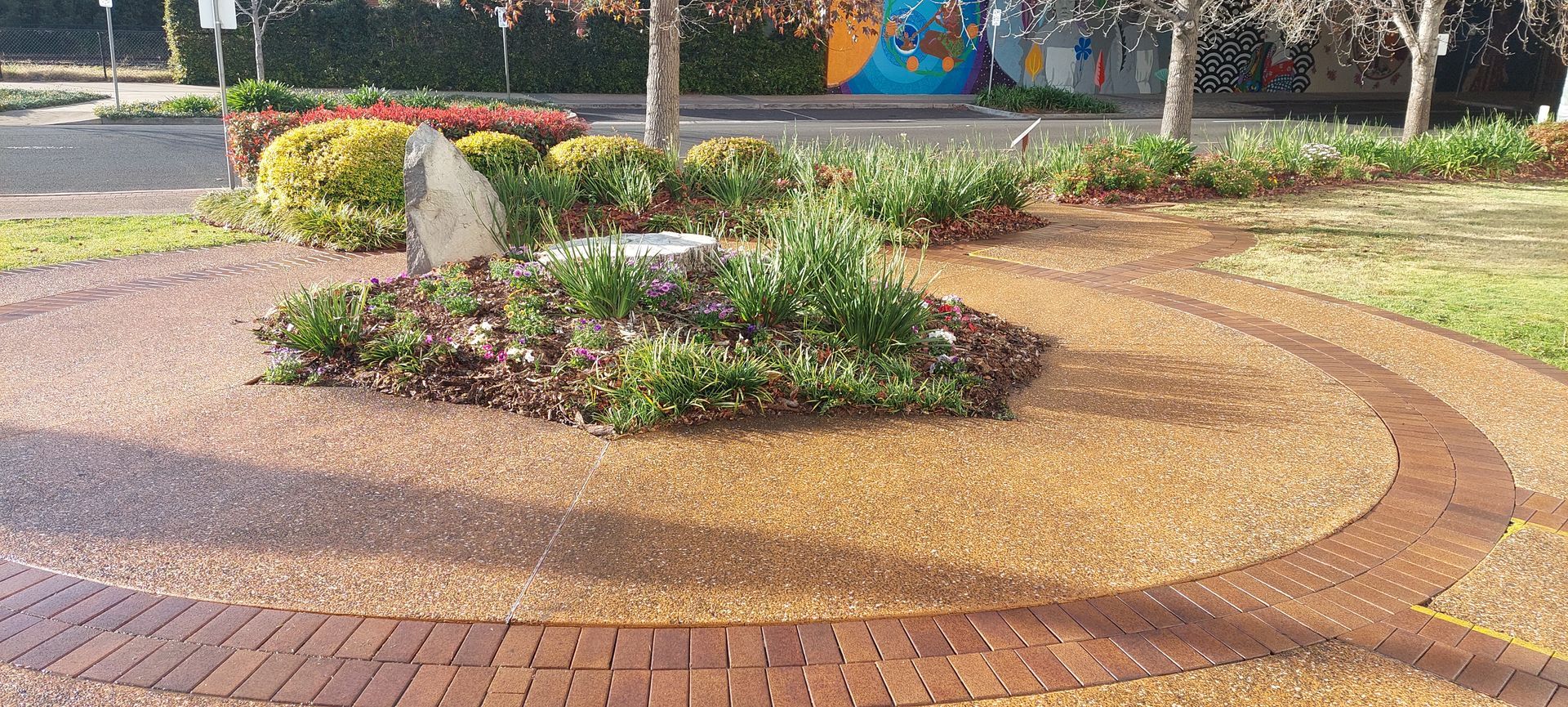 A circular garden bed featuring a large stone and various shrubs, surrounded by decorative gravel and a brick border.