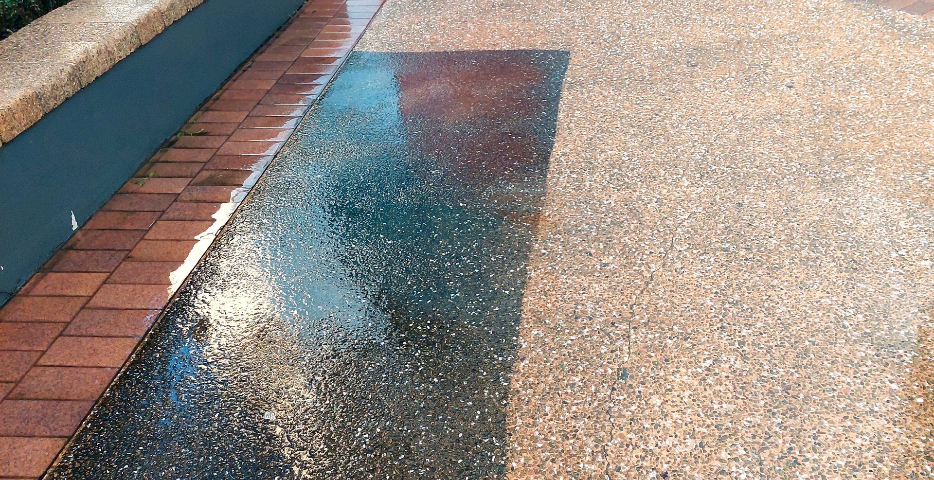A puddle reflects a building on a pebbled concrete walkway next to a row of reddish-brown bricks.