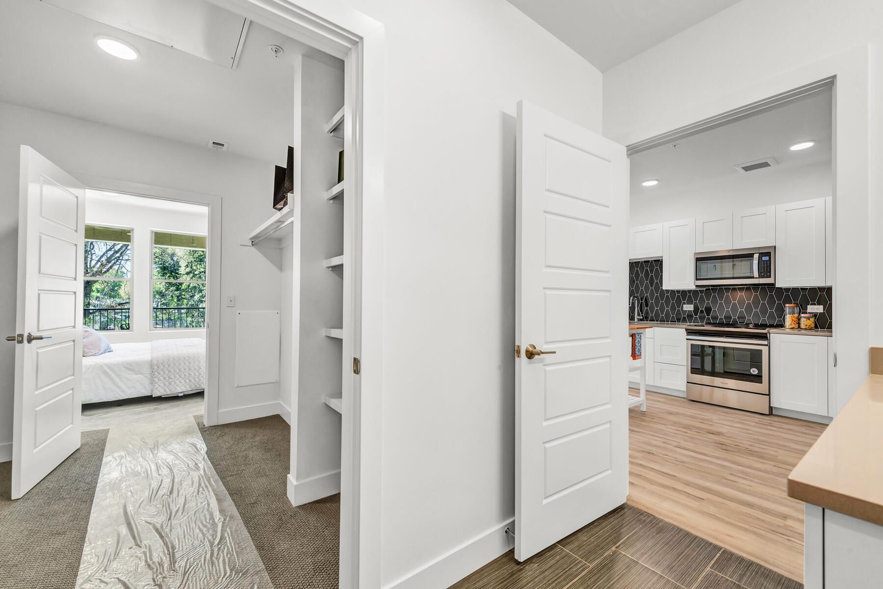 Interior hallway with open doors to a bedroom, kitchen, and closet. White walls, wooden floors, and natural light.