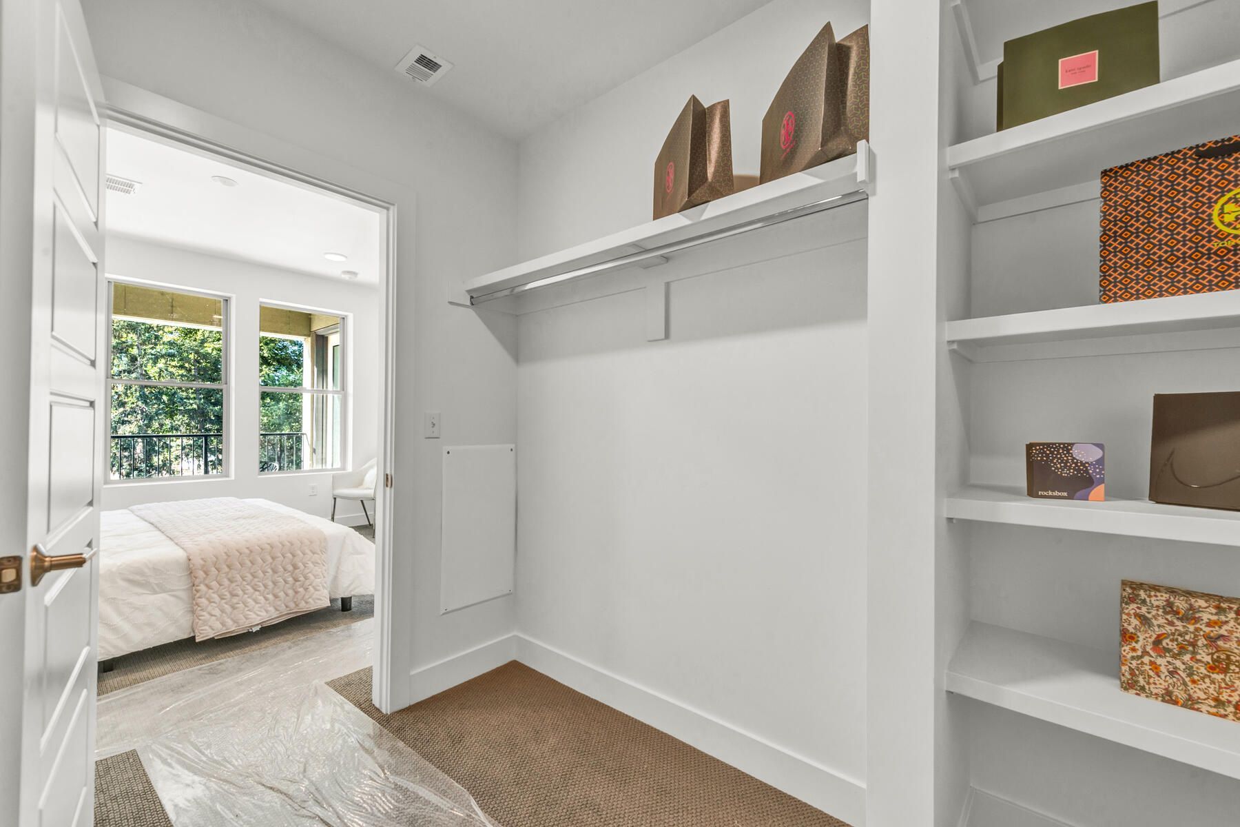 Walk-in closet with shelves and rod. View into bedroom with bed. White walls, carpet.