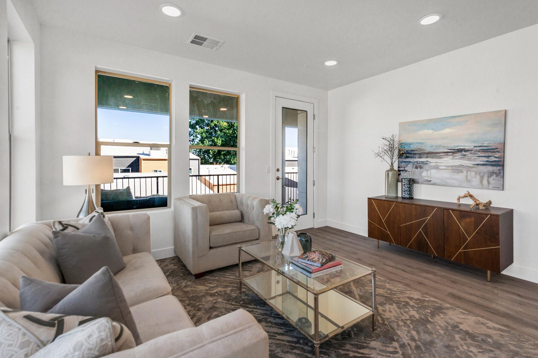 Living room with a sofa, armchair, coffee table, rug, artwork, and wood cabinet, next to windows with outdoor views.