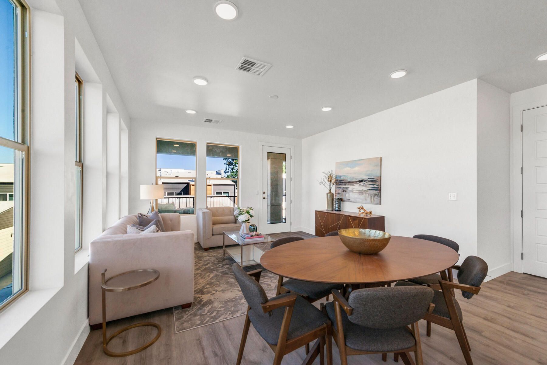 Bright, modern living room with dining table, sofa, and windows. White walls, wood floor, neutral colors.