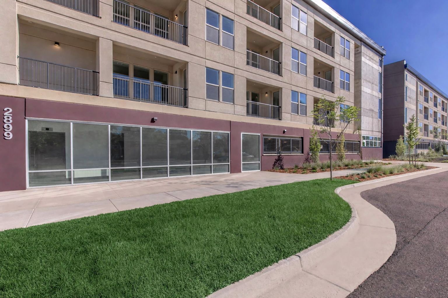 Multi-story beige apartment building with commercial space. Green lawn, sidewalk, and street.