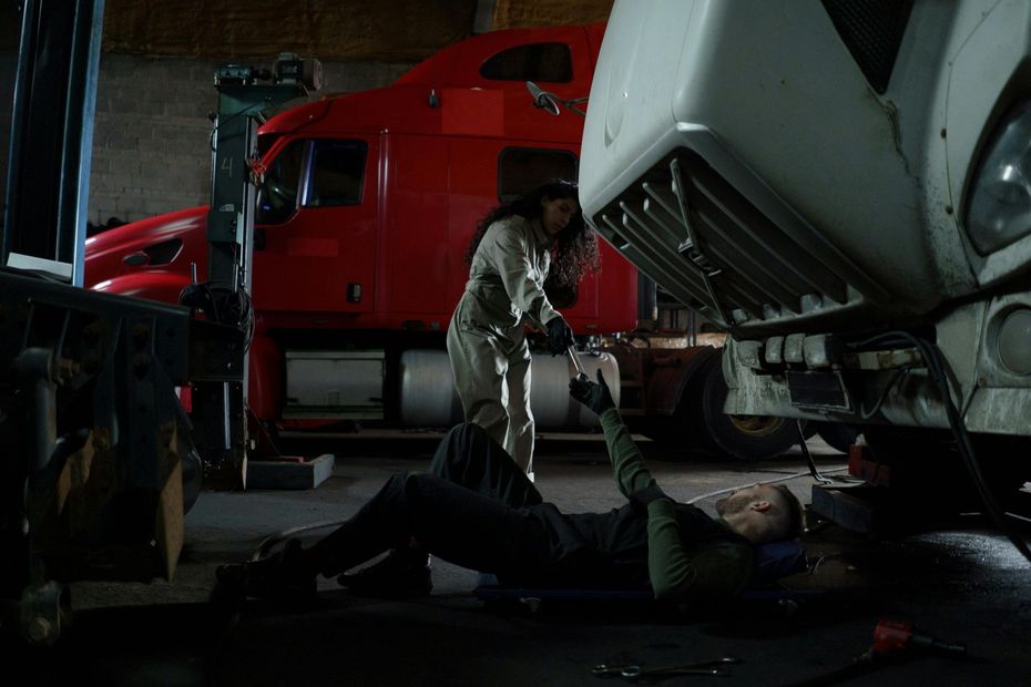Two people working on truck in garage; one under truck, one standing. Red and white truck.