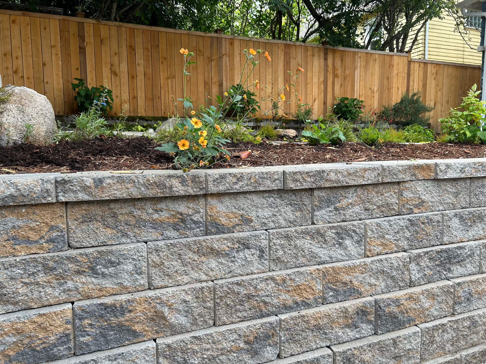 A stone wall with a wooden fence in the background and flowers in the foreground.