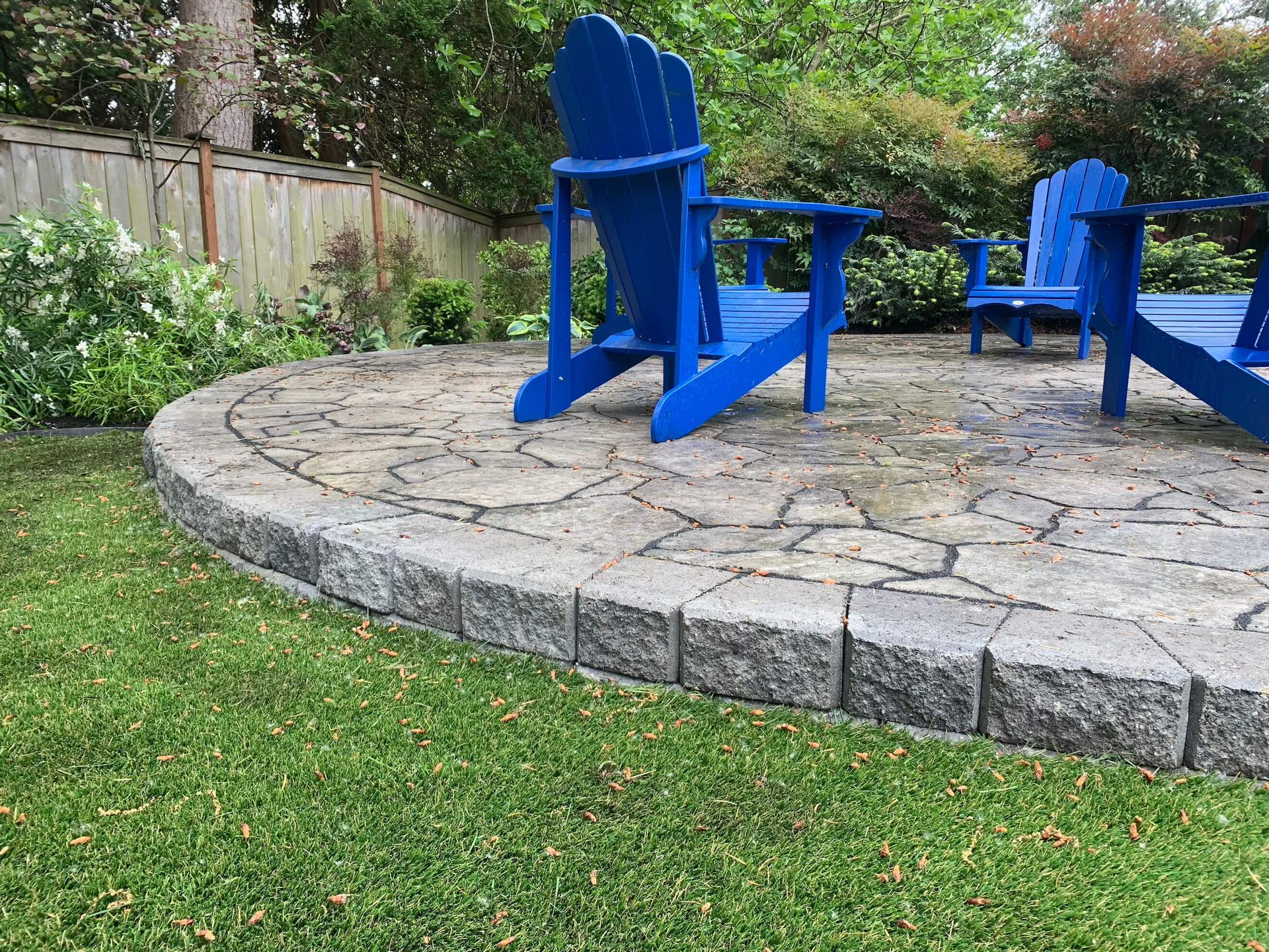 Two blue adirondack chairs are sitting on a stone patio in a backyard.