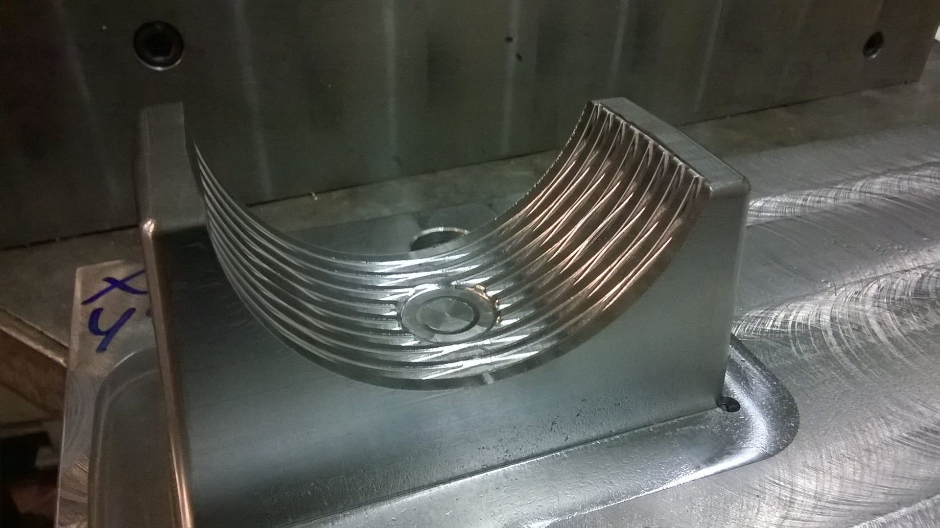 Un pezzo metallico lavorato con creste curve, posizionato su una base metallica, con un foro e la marcatura XY.