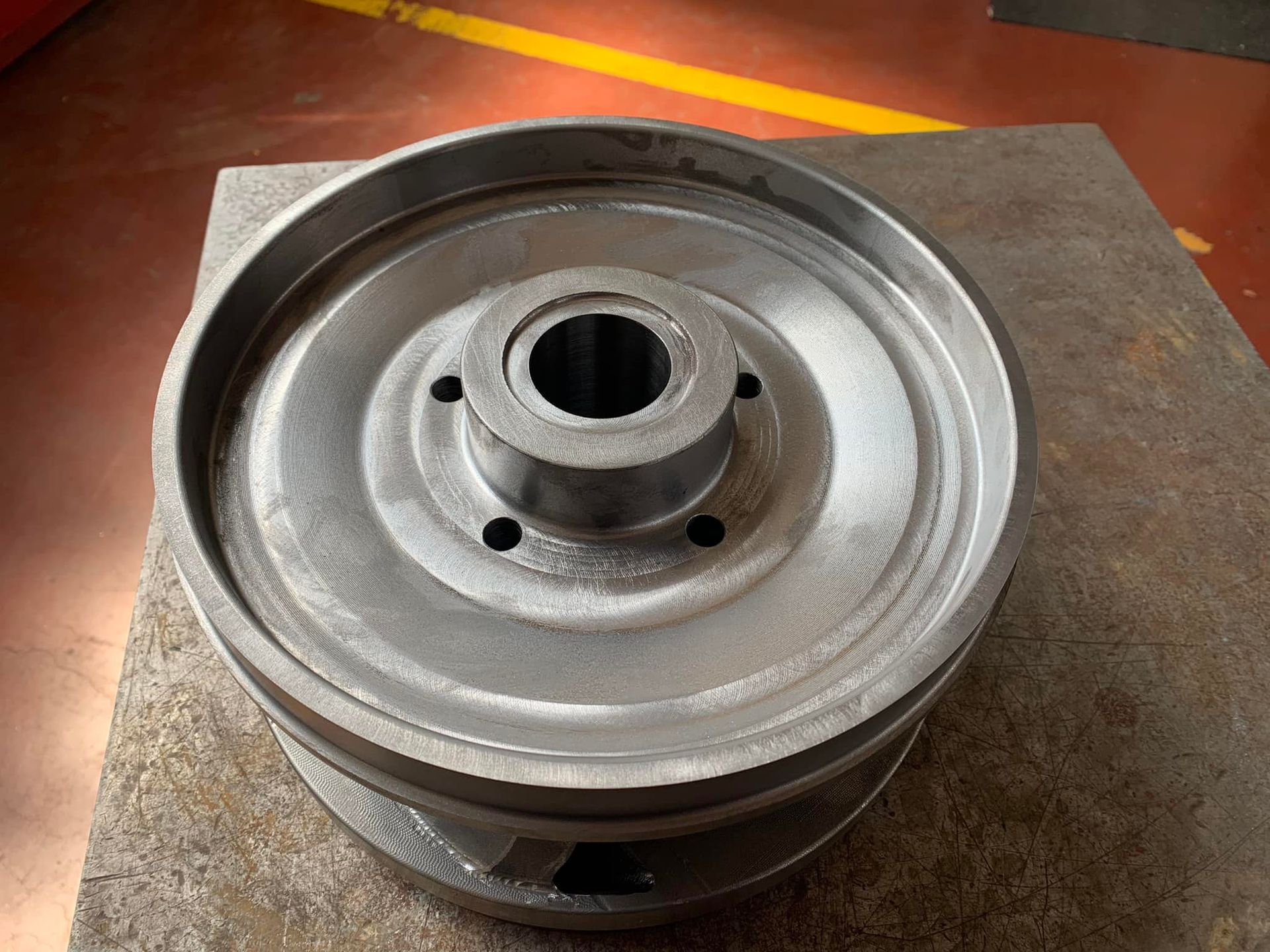 Puleggia in metallo grigio con mozzo centrale e fori per bulloni, appoggiata su una superficie metallica.