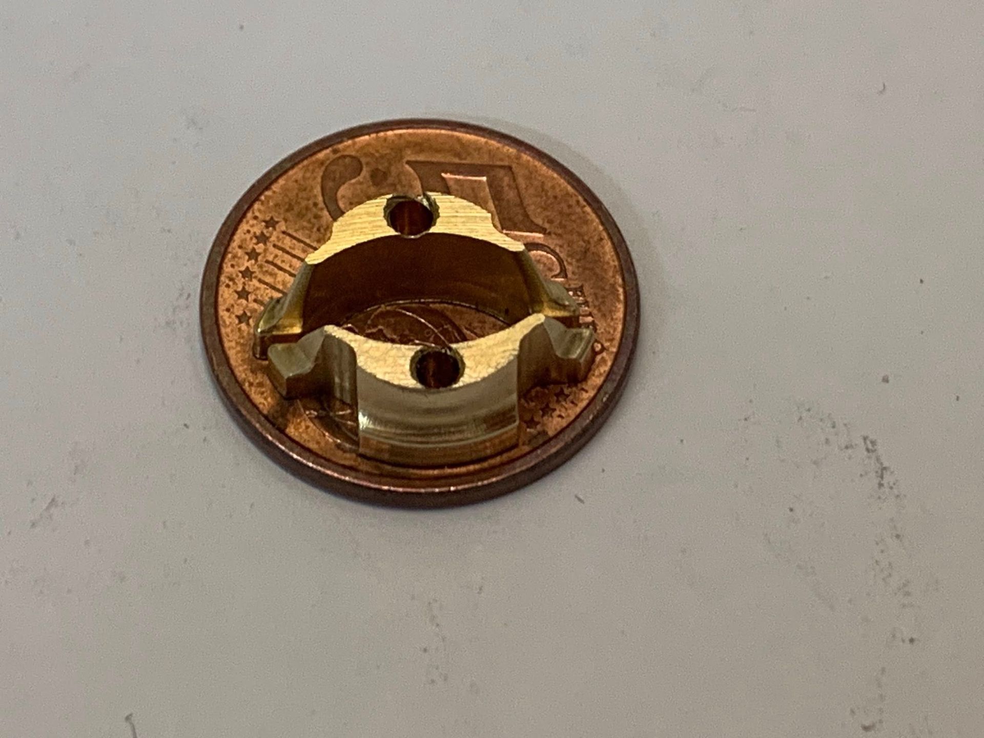 Pezzo metallico circolare color ottone con tre fori, appoggiato su una moneta di rame.