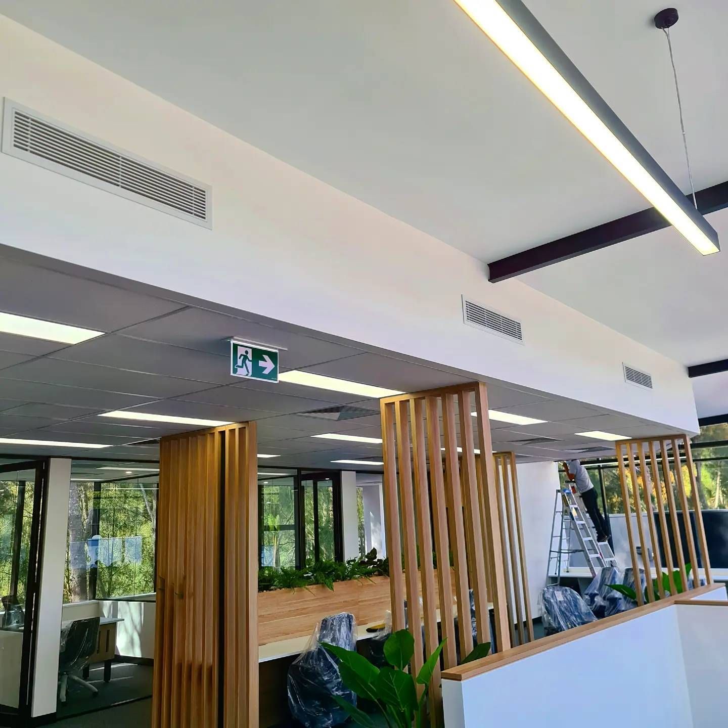 A man is standing on a ladder in a room with a green exit sign on the wall. — Coastline Air In Forresters Beach, NSW