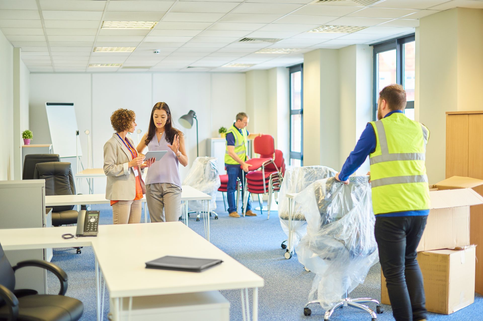 Office relocation: Workers move chairs and boxes while two people converse in a bright, modern office space.