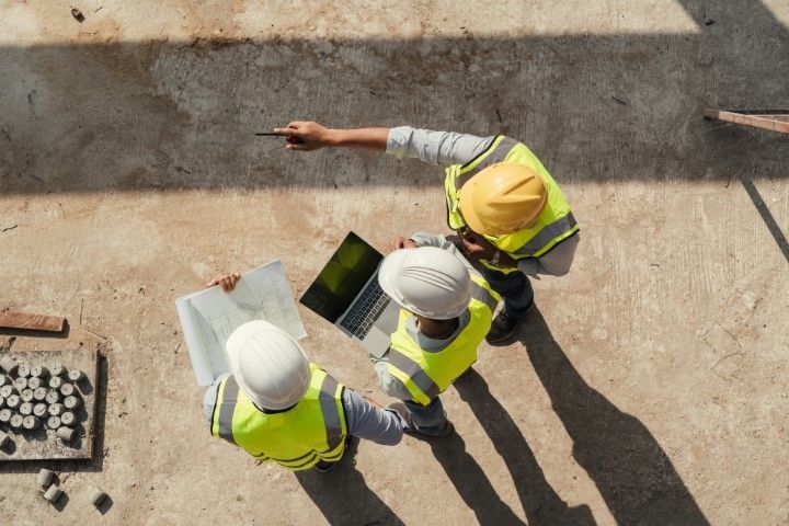 Three construction workers in hard hats and safety vests consult blueprints and a laptop on a job site.