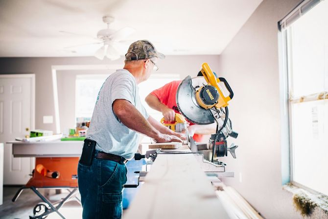 A person in a light shirt and camouflage hat uses a miter saw to cut a wooden board in a room under construction.