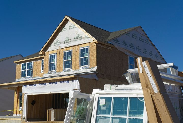 A two-story residential house under construction, with exposed wood framing, protective house wrap, and new windows onsite.