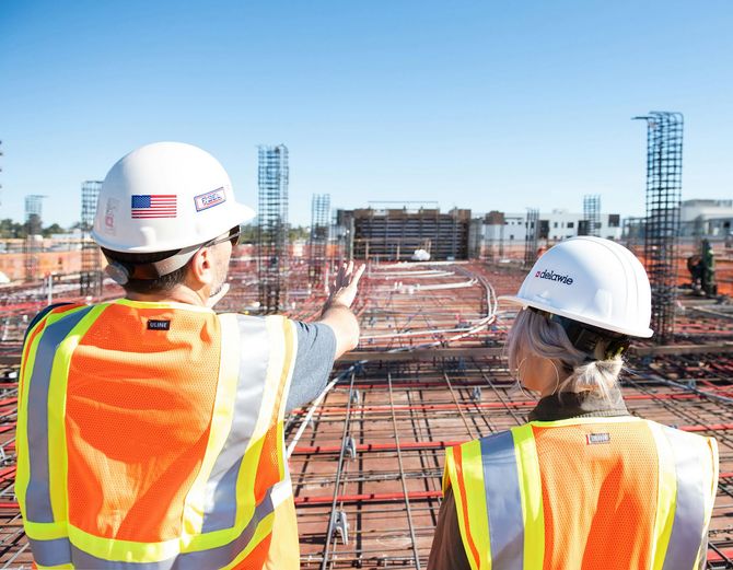 Two construction workers in hard hats and safety vests stand on a concrete site, observing building framework.