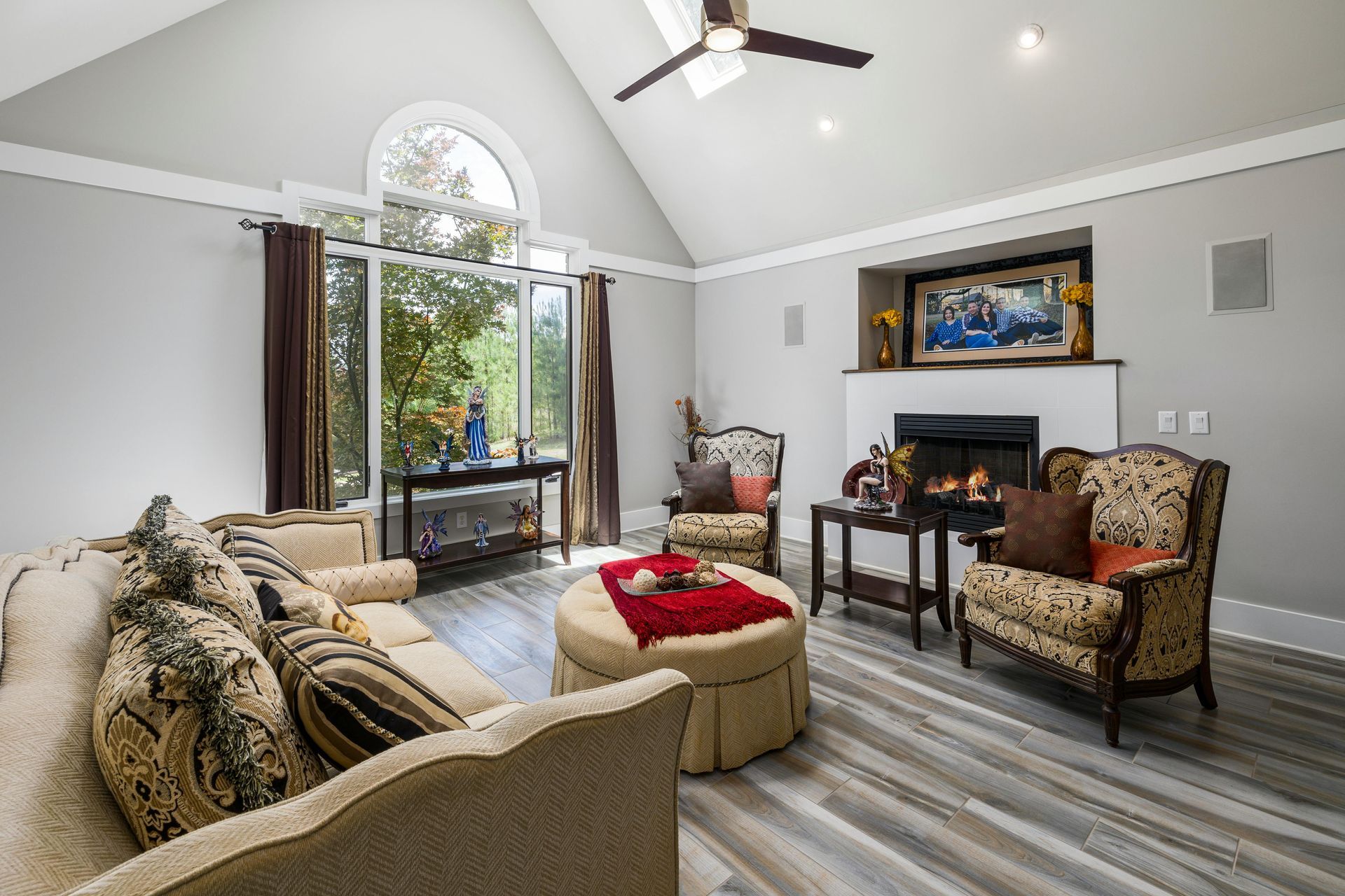 A cozy living room with a fireplace, patterned armchairs, a couch, and a large arched window under a vaulted ceiling.