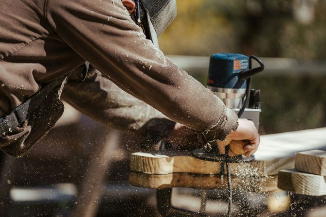 A person using a handheld power router to shape the edge of a wooden board, with wood shavings flying in the air.