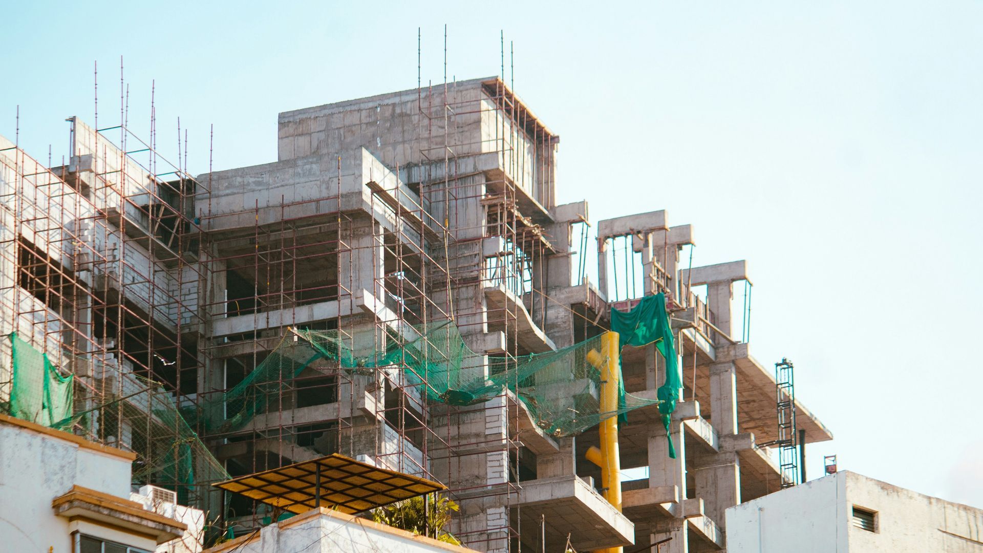 A tall concrete building under construction, surrounded by metal scaffolding and protective green safety netting.