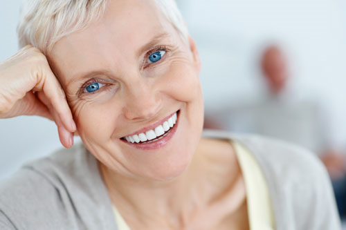 An older woman with blue eyes is smiling with her hand on her head.
