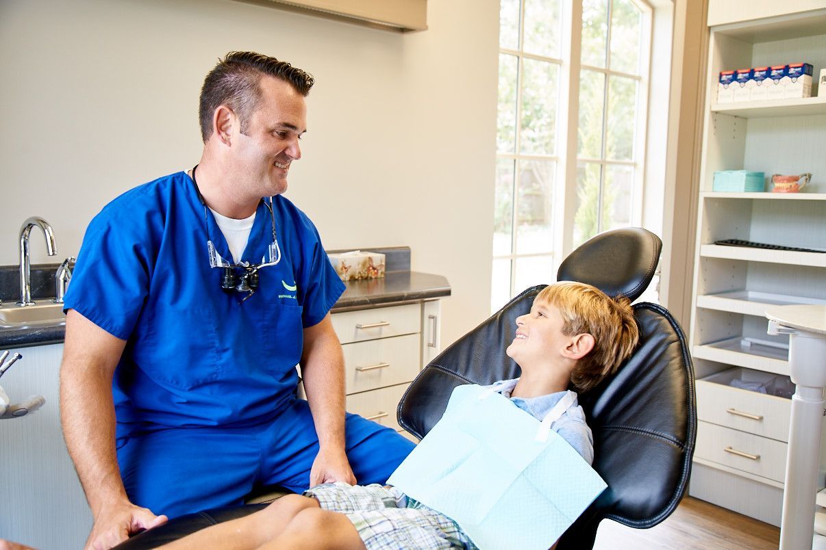 Dentist in blue scrubs talking to a young boy in a dental chair. Sunlight streams in a window.