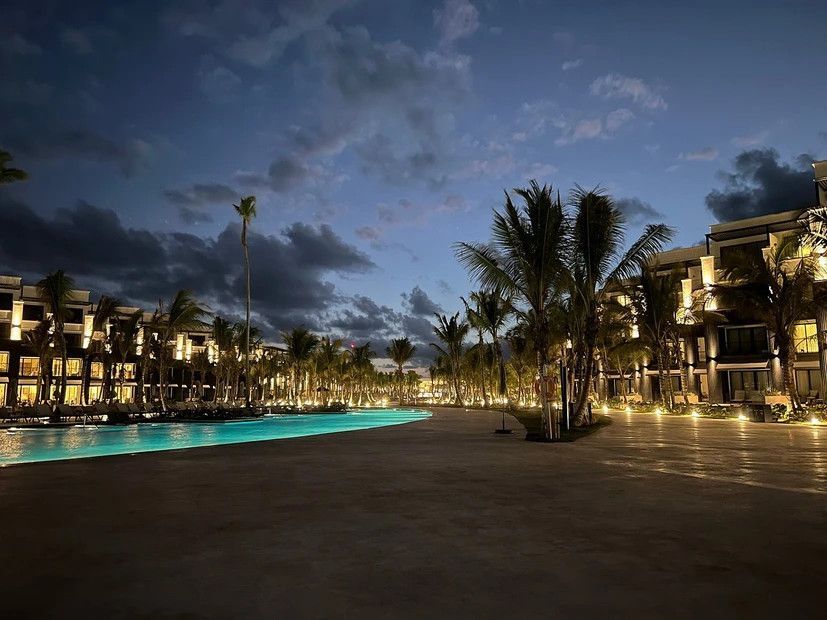 A large swimming pool is surrounded by palm trees and buildings at night.